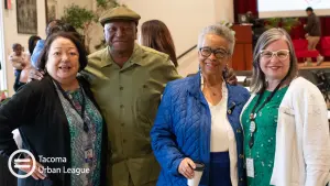Four Tacoma Urban League community members smile together at an indoor gathering: an older woman in a black cardigan, a man in a green shirt and flat cap, a woman in a blue quilted jacket, and a woman in a green patterned blouse with a white cardigan; the Tacoma Urban League logo watermark appears at lower left